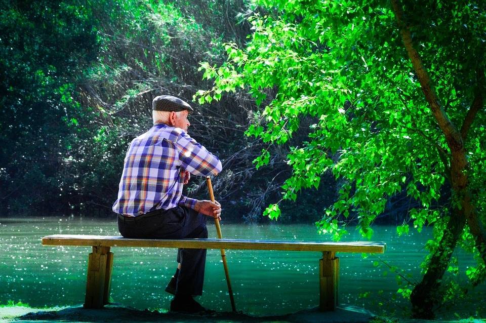 Fotografía de un hombre de avanzada edad, con bastón y boina, sentado en un banco frente a un estanque, en un parque.