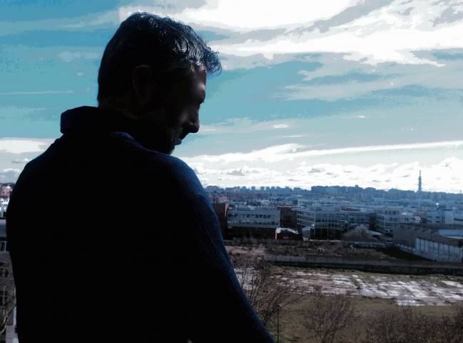 Fotomontaje titulado "La soledad". La autora es Laura Rico. Se ve de fondo un paisaje urbano frío y azulado. Y en primer término, de espaldas, la silueta a contraluz de un hombre que mira al suelo con aspecto triste. 