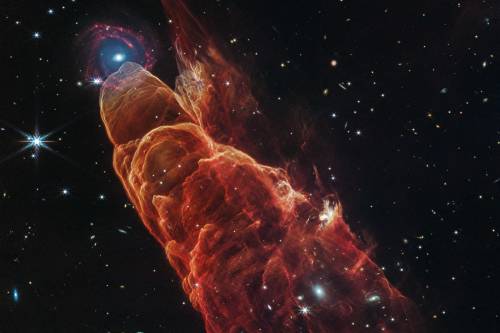La nube de forma cónica de color rojo anaranjado corresponde a Herbig-Haro 40/50. En el extremo superior izquierdo hay una galaxia de fondo en forma de espiral con un centro azul.