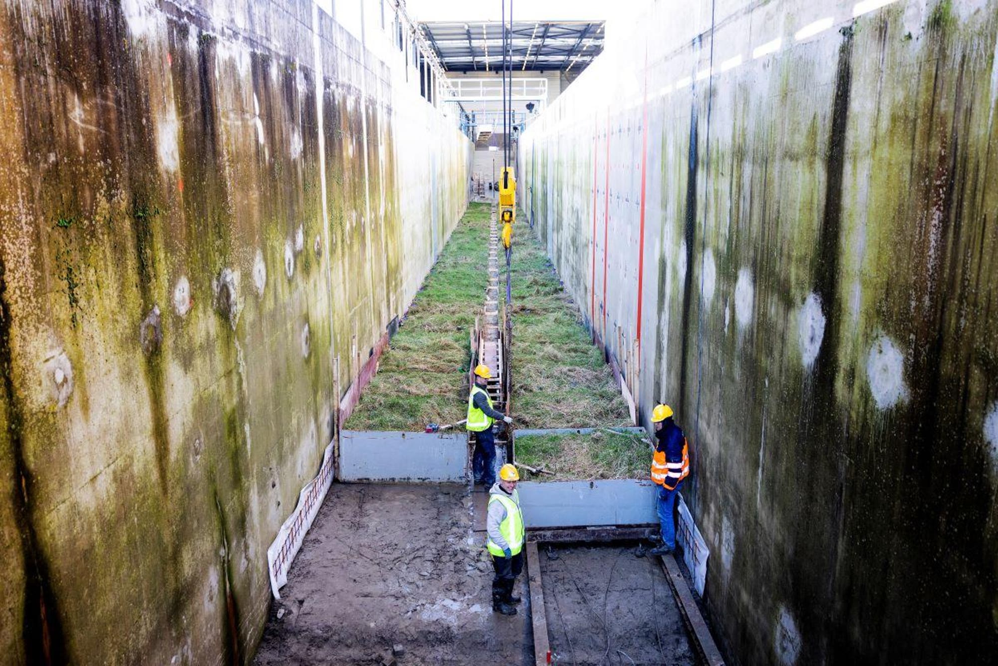 Operarios con casco y chaleco reflectante trabajan entre las altas paredes de hormigón de un canal, sobre bloques cubiertos de hierba.