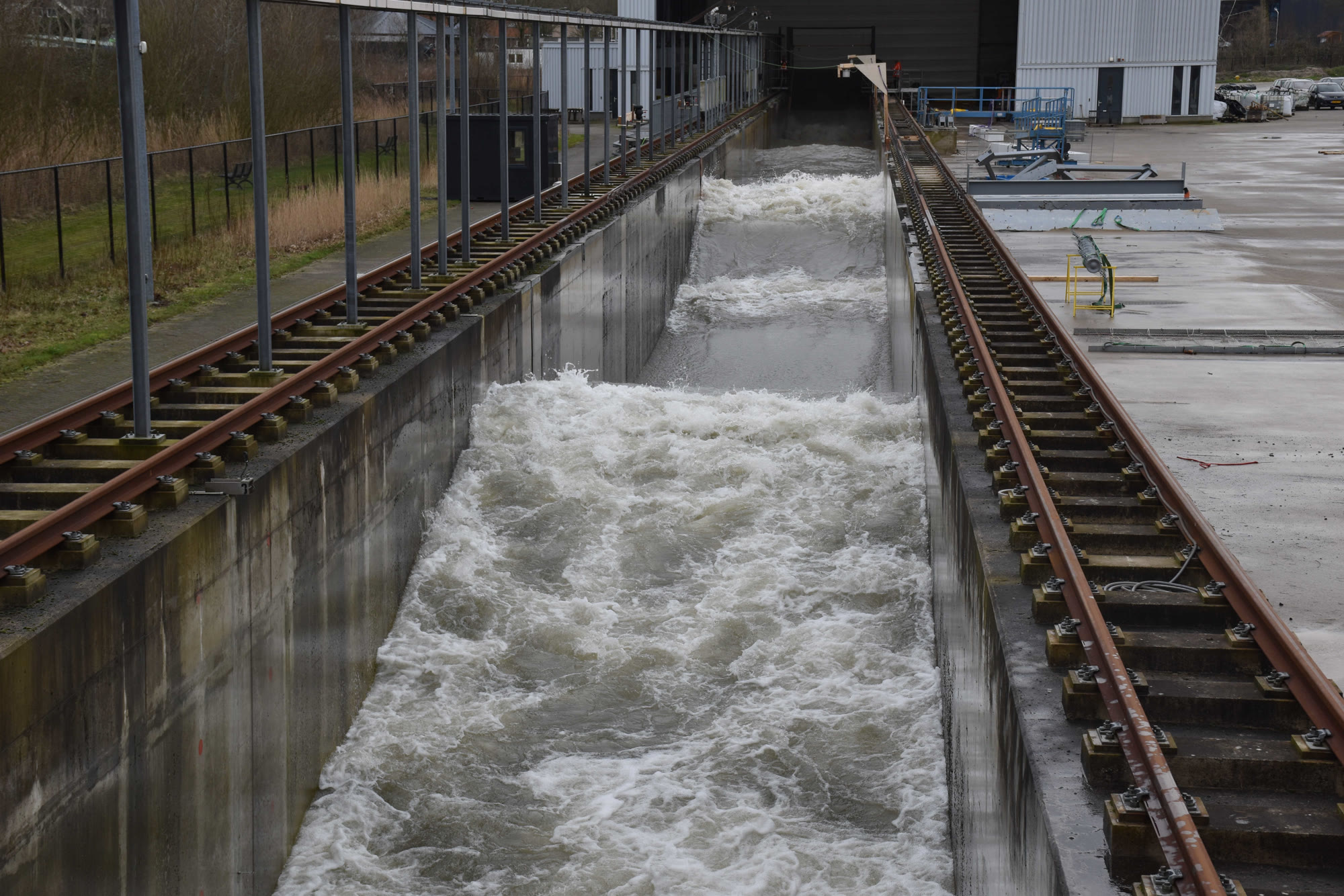 Olas cubiertas de espuma avanzan por un largo canal de hormigón flanqueado por raíles.
